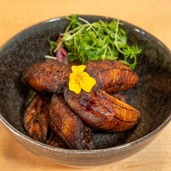 Manjay Restaurant's caramelized plantains with a yellow edible flower and fresh greens in a dark bowl.