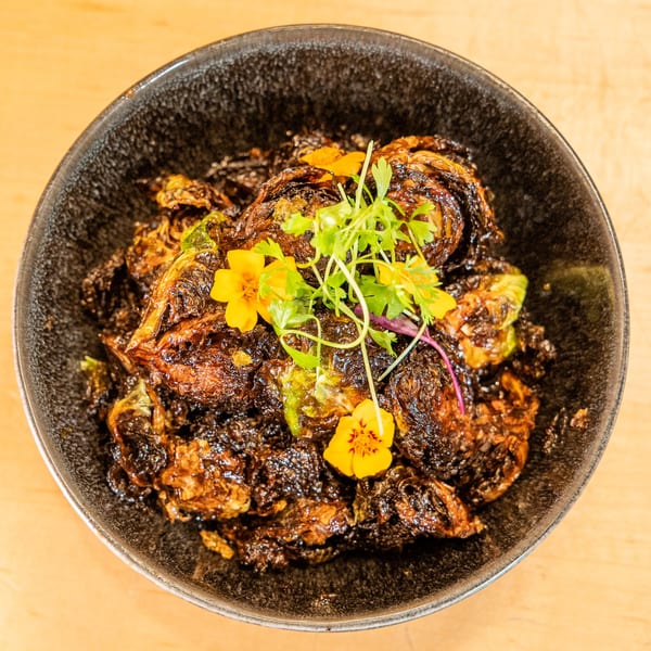 Crispy, dark-fried brussels sprouts garnished with yellow edible flowers and fresh herbs in a black bowl.