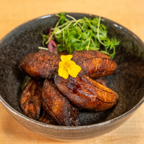 A black bowl filled with caramelized plantains, garnished with microgreens and a yellow edible flower.