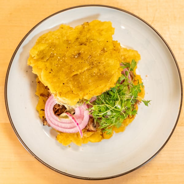 A Caribbean sandwich on a white plate, featuring plantain buns, pulled pork, red onions, and microgreens.